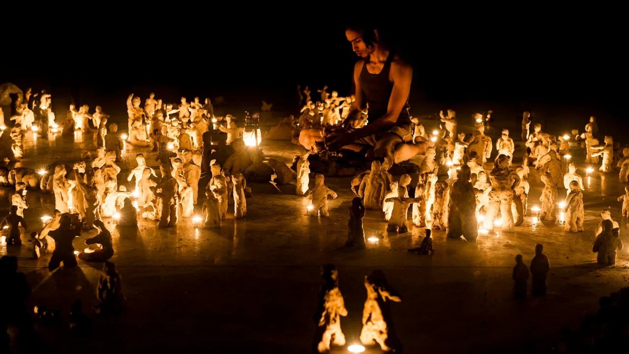 Artist sits on the floor in a dar room, surrounded by candles and clay figures
