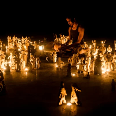 Artist sits on the floor in a dar room, surrounded by candles and clay figures