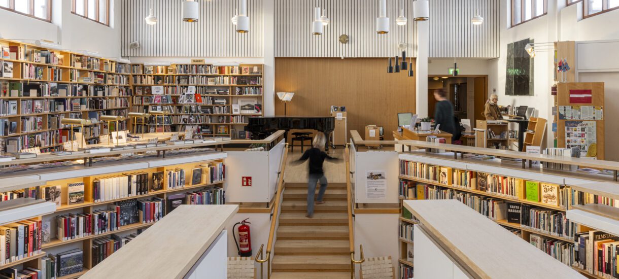Image shows the Nordic House library. A staircase is in the middle, a child running down the stairs. All around are bookshelves filled with books. Big windows above the shelves and the daylight coming from them lights up the spaces.