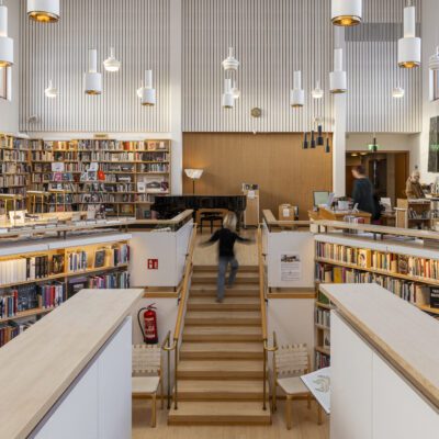 Image shows the Nordic House library. A staircase is in the middle, a child running down the stairs. All around are bookshelves filled with books. Big windows above the shelves and the daylight coming from them lights up the spaces.