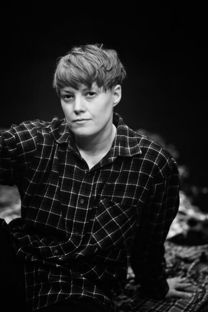 Black and white photo of a woman, Ann-Christin Kongsness, sitting down and looking straight into the camera. She wears a plaid shirt and has short hair. 