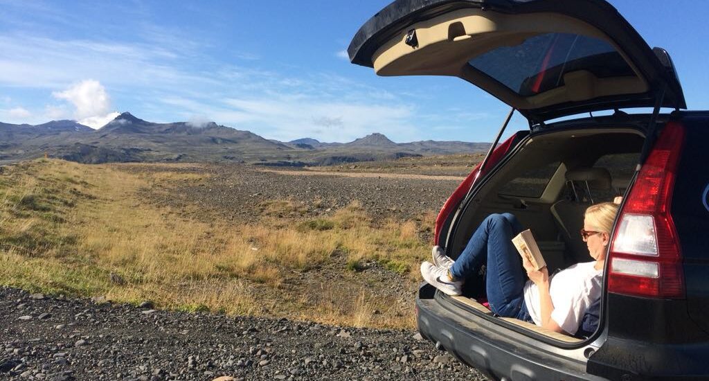blond woman sits in a open car trunk and reads a book, landscape of lava and mountains