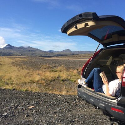 blond woman sits in a open car trunk and reads a book, landscape of lava and mountains