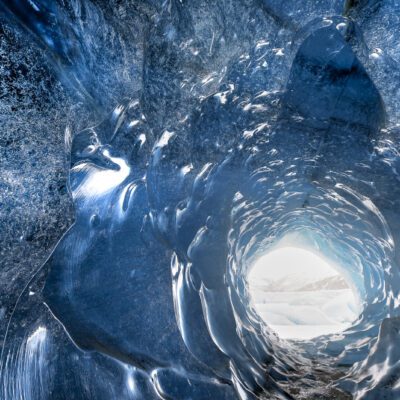 A photograph taken inside a glacier cave, at the end of the cave you see an opening. The image is mostly blue in color.