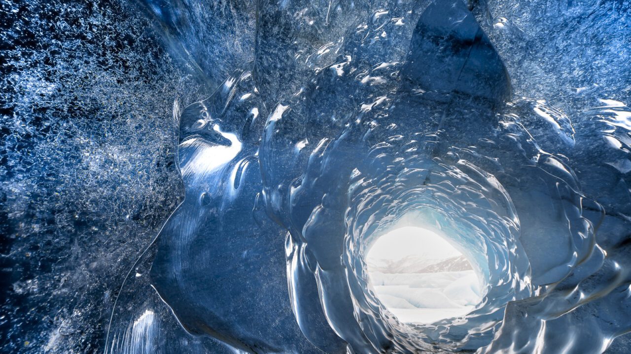 A photograph of a glacier cave, blue color and a bright white opening at the end of the cave