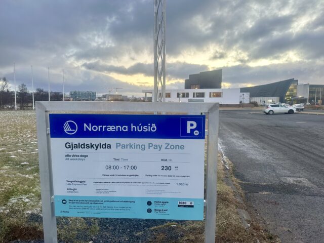 Photograph of a blue, white and teal colored aluminum sign outside a parking lot. On the sign is a text describing parking information. 
