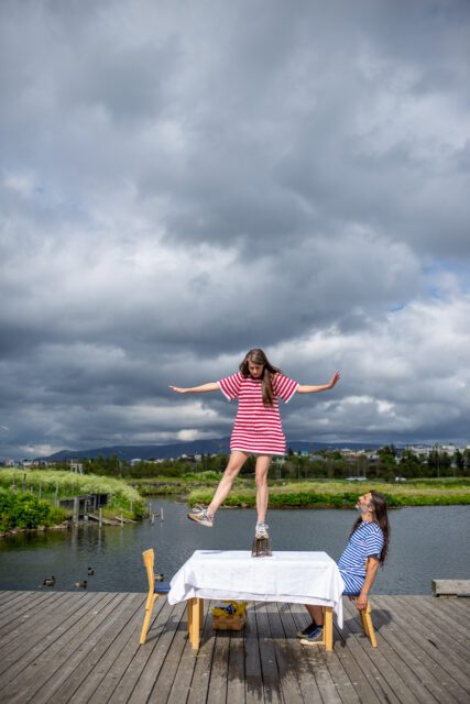 From an art performance. A woman stands on top of a alvar aalto vase on top of a table. A man sits at the table and watches her. Nature in the background