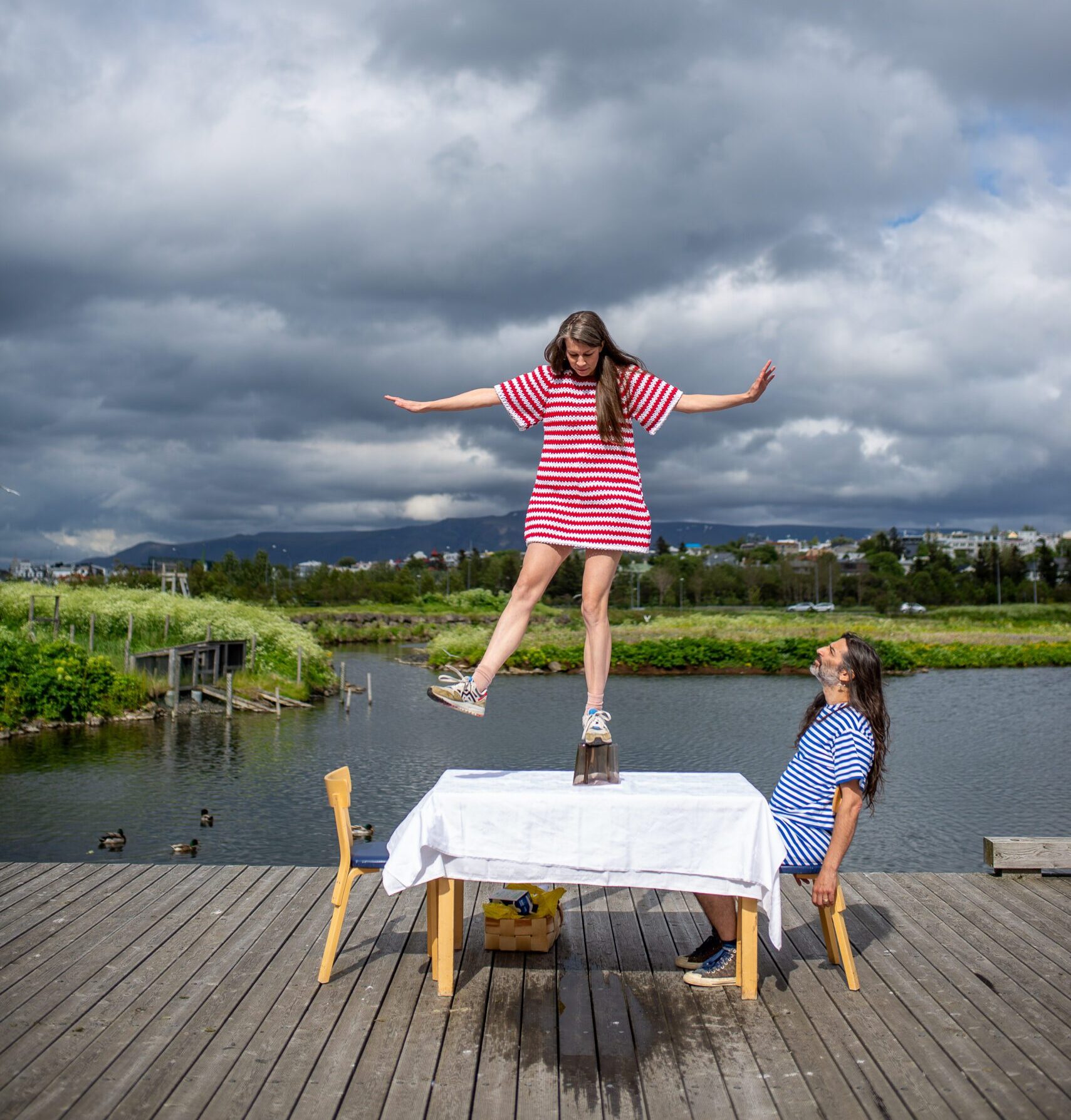 From an art performance. A woman stands on top of a alvar aalto vase on top of a table. A man sits at the table and watches her. Nature in the background