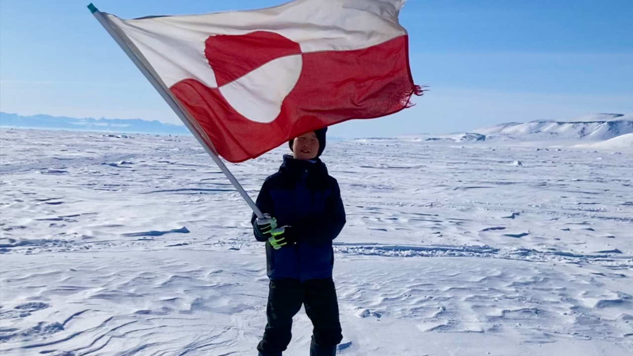 A boy holding a Greenlandic flag standing on snow with blue skies in the background.
