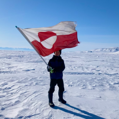 A boy holding a Greenlandic flag standing on snow with blue skies in the background.