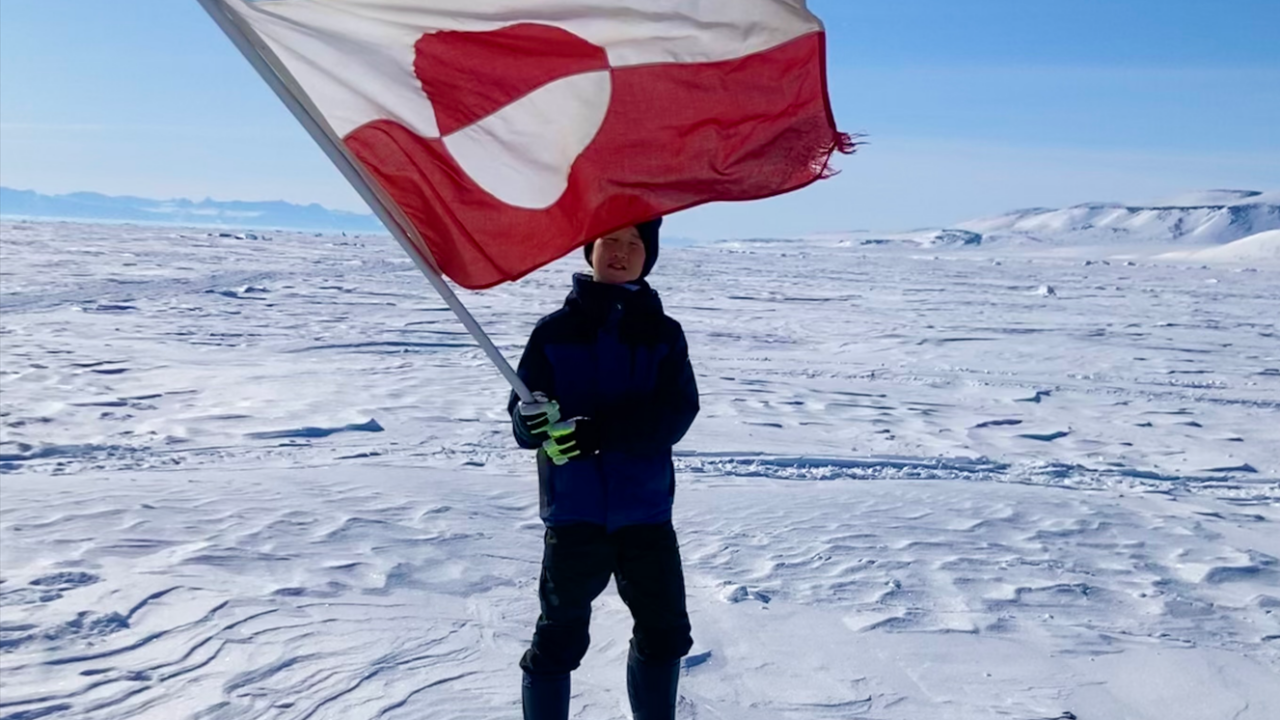 A boy holding a Greenlandic flag standing on snow with blue skies in the background.