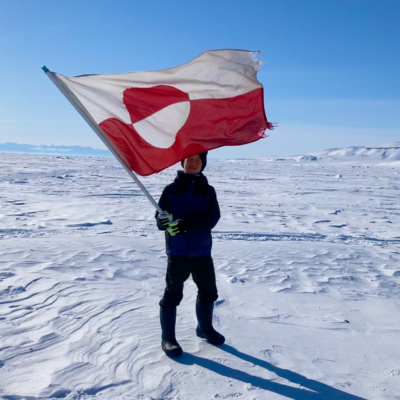A boy holding a Greenlandic flag standing on snow with blue skies in the background.