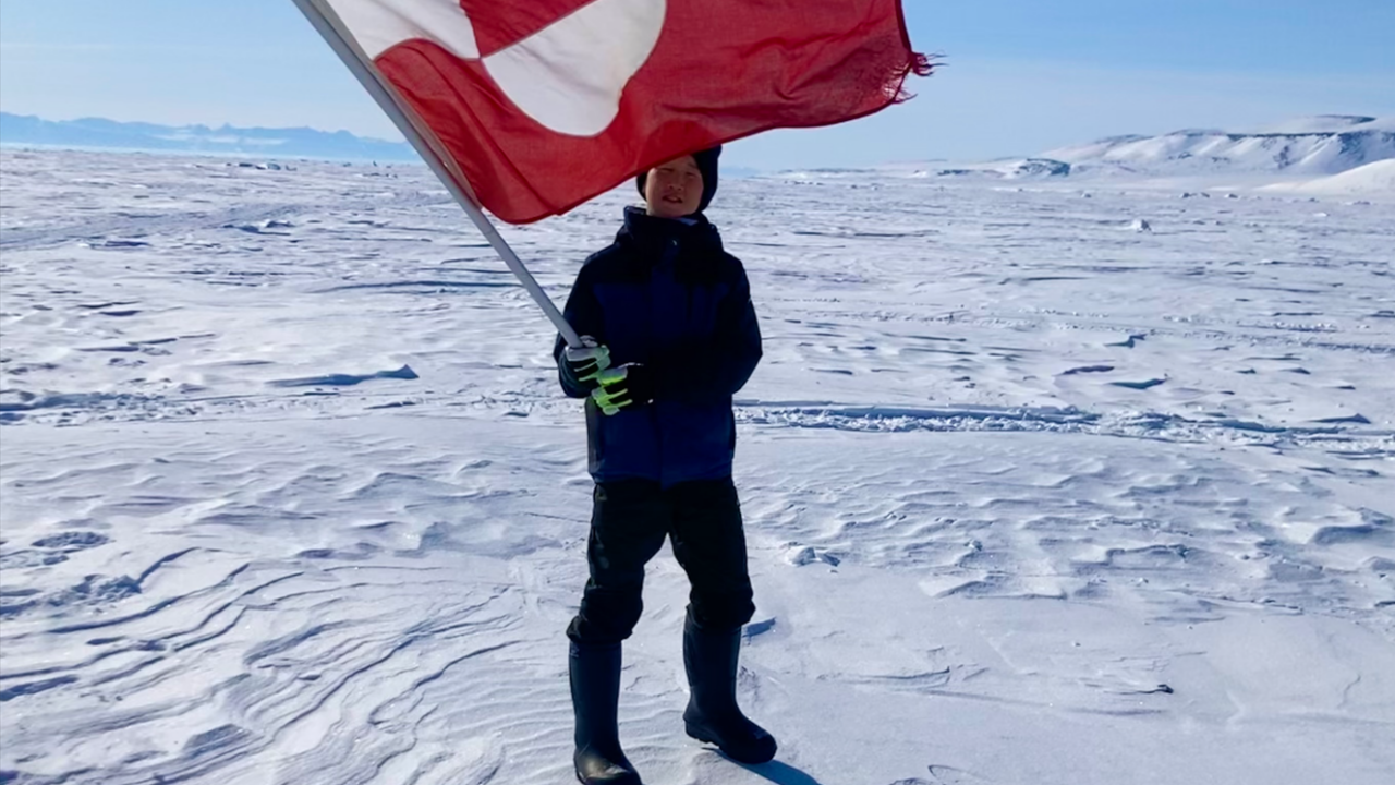 A boy holding a Greenlandic flag standing on snow with blue skies in the background.