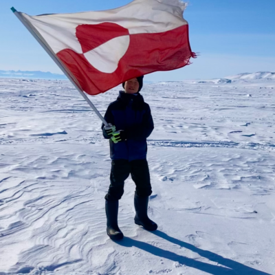 A boy holding a Greenlandic flag standing on snow with blue skies in the background.