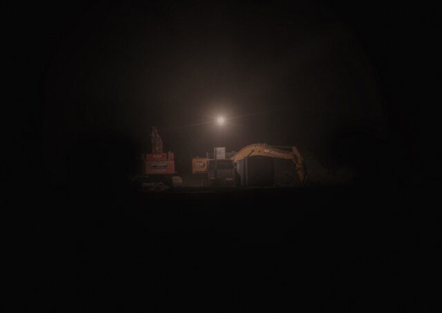 Photo taken at night, very dark and in the distance you can see a construction machine under a small light.