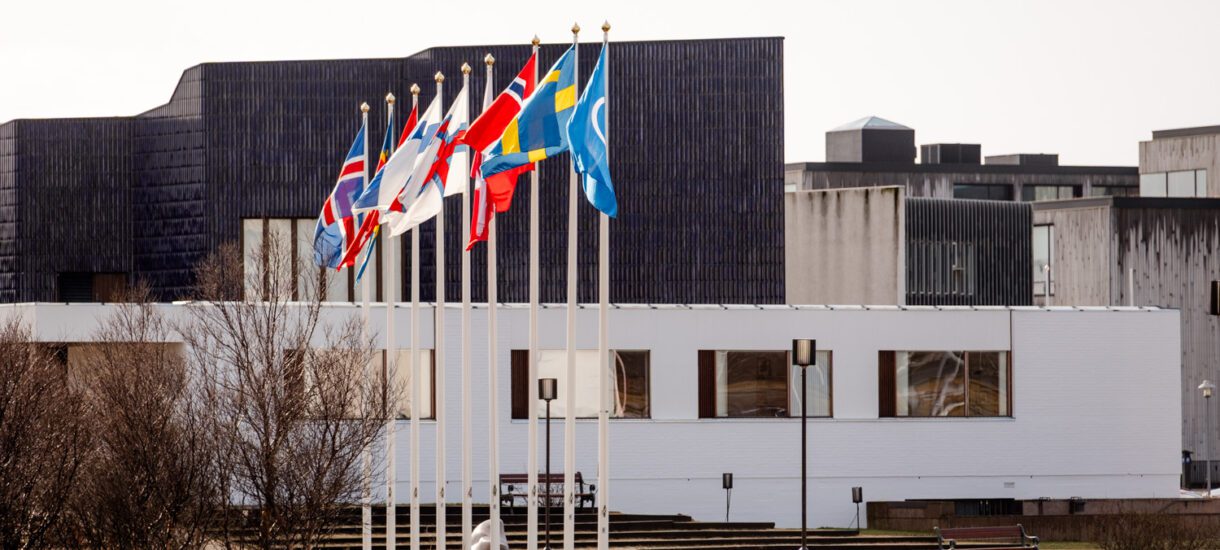 A photograph showing nine flags from various Nordic countries flying on poles, set in front of a modern white building with a blue roof.