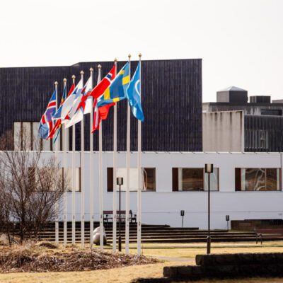 A photograph showing nine flags from various Nordic countries flying on poles, set in front of a modern white building with a blue roof.