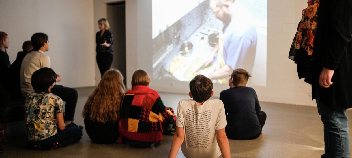 Children sitting on the floor listening to a guided tour of an exhibition. A video work on the wall in front of them. A persion stands infront of the work and seems to be guiding them. The picture is taking from behind the kids.