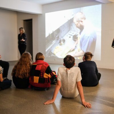 Children sitting on the floor listening to a guided tour of an exhibition. A video work on the wall in front of them. A persion stands infront of the work and seems to be guiding them. The picture is taking from behind the kids.