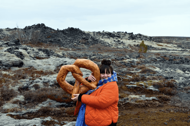 Artist Estelle Pollaert standing in a lava field. Hugging a giant paper maché pretzel. She is wearing an orange coat and blue skarf. She has dark brown hair.