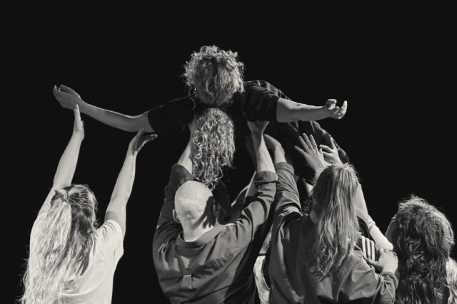 Black and White image of people dancing. They all turn their back to the camera, four people lift one person up above their heads.