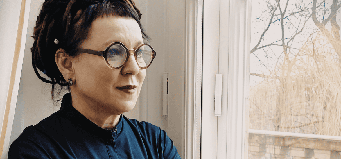 photo of a woman standing by a window and looking out. She has glasses and wears blue.