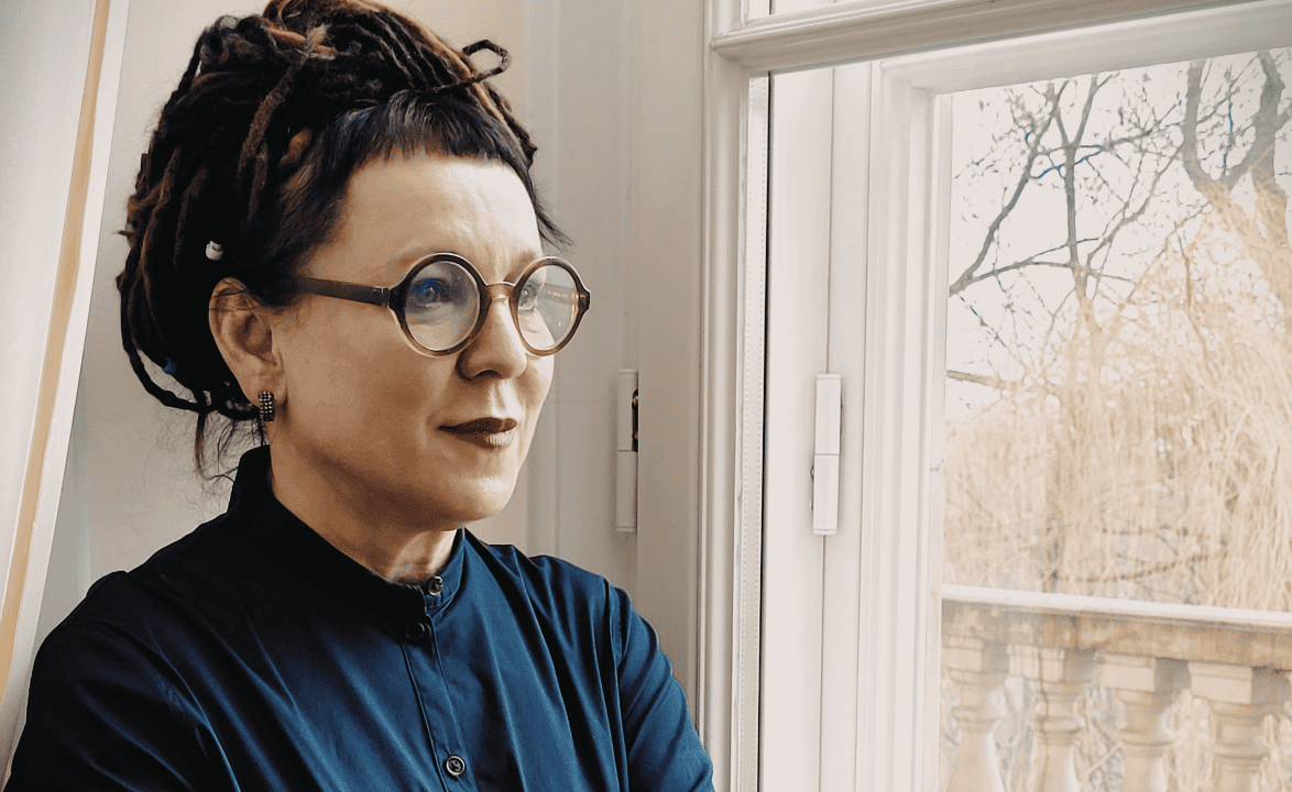 photo of a woman standing by a window and looking out. She has glasses and wears blue.