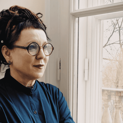 photo of a woman standing by a window and looking out. She has glasses and wears blue.
