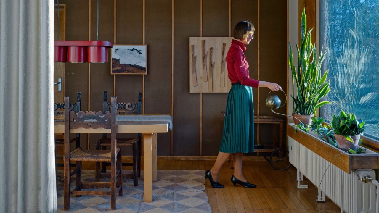 A woman wearing red shirt and blue skirt stands by a window and is watering plants. Light falls on her face.