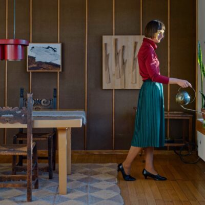 A woman wearing red shirt and blue skirt stands by a window and is watering plants. Light falls on her face.