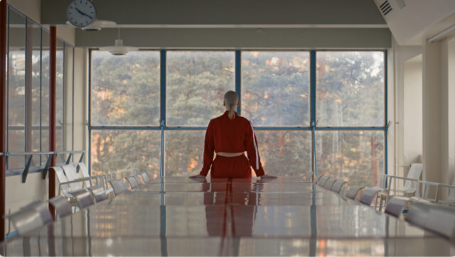 A person leans to a table, back to camera, facing a large window. The person wears a red tracksuit.