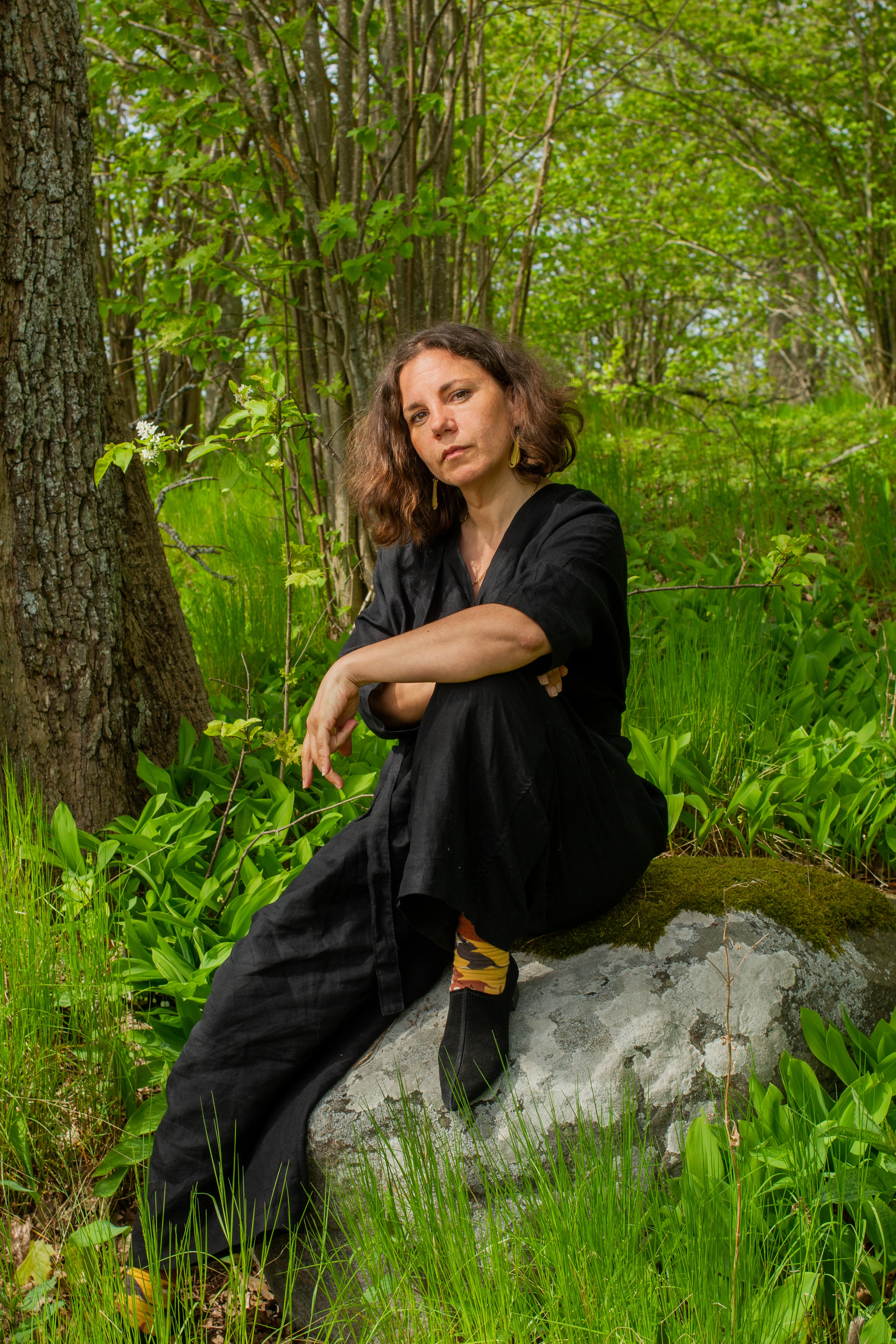 portrait of a woman with brown hair, she sits on a rock, surrounded by nature 